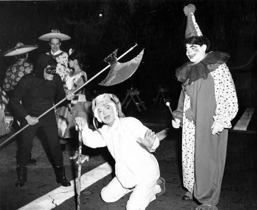 A person in white kneeling, another in clown costume, and another as an executioner