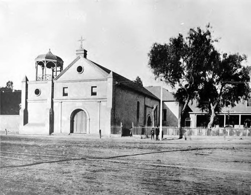 View of Plaza Church with dirt road in foreground and two men to the right of the church