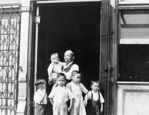 Elderly man standing in storefront doorway holding a baby with four young boys standing in front of him at 425 N. Los Angeles Street, Howard Quon is the second from the left