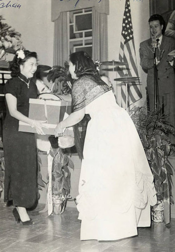 Lily Chan, chair of the women's division at the Chan Association on Bernard Street in Los Angeles, presenting gifts to Ms. Doris Chan of Hong Kong (later Mrs. Mok). She is the daughter of famous Chan Ng Mun Chee (educator) of Hong Kong. Tien Son Chan is on the stage