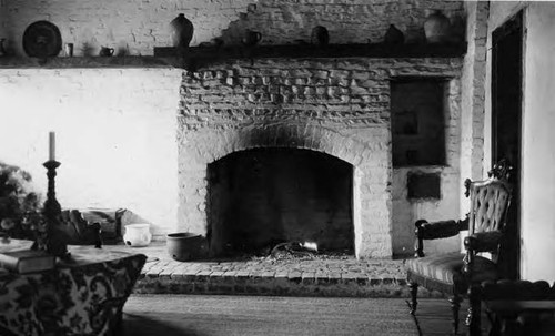 Interior shot of Avila adobe showing fireplace with small flames in grate. Assorted pottery on mantel and on floor near fireplace. Bench against fireplace wall; chair against door; area rug on wooden floor; table with candlestick and tablecloth and book shown