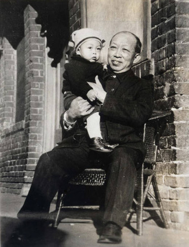 Elderly man sitting in a chair holding a baby, the uncle of Howard Quon