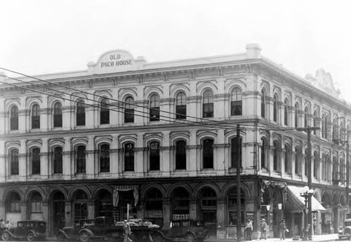 Pico House, pediment says "Old Pico House." Awning on west facade, cars and people on west and north sides, old lamppost on west side, sidewalk in front, with telephone pole and wires across front