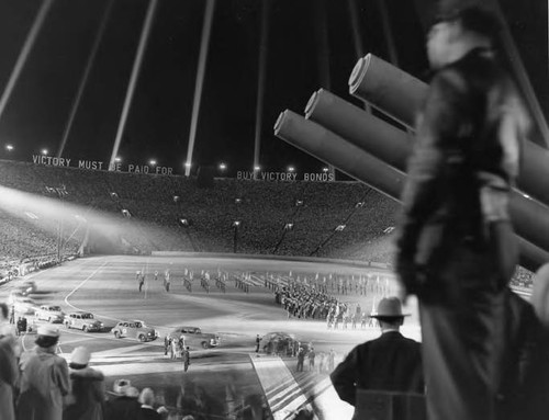 A large stadium celebration in support of the war effort. Large letters at the far top edge of the stadium read: "VICTORY MUST BE PAID FOR. BUY VICTORY BONDS." Three canons protrude from the right side and a line of cars enter from the left. Troops assemble on the field and the stadium is packed with supporters