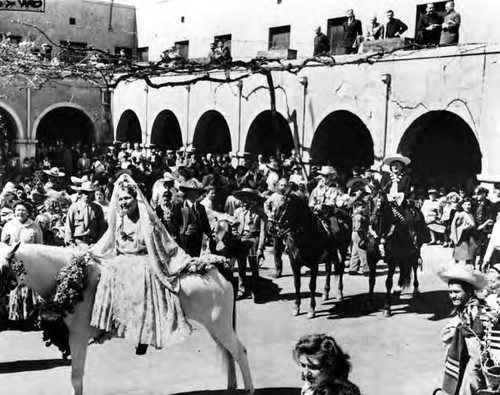 Crowds of people marching in a parade, standing around as if posed for pictures. Christine Sterling is in the photograph, smiling while sitting on a horse