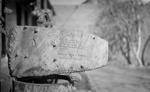 Avila adobe in background (fuzzy). On a wood cistern containing water (a pump handle appears to be attached, a wood sign that says "From the willow tree where Allesandro first saw RAMONA as she was washing the altar cloth at the river. Camulos- home of Ramona, 18 mils west of Newhall."