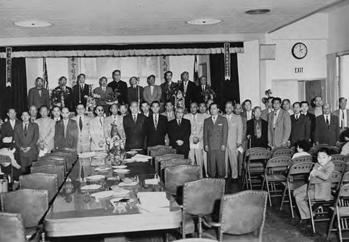 Posed photo of a large group of Chinese men in the Chinese Consolidated Benevolent Association Assembly Hall, obviously a formal occasion; pictured among the prominent leaders on the stage is Gee Kee Ward; taken in Los Angeles