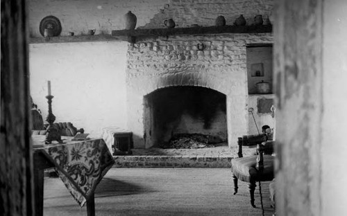 Interior shot of Avila adobe showing fireplace with brazier and andiron against fireplace. Assorted pottery on mantel and in niche. Two chairs and a table with a candle stick on it are also shown. Area rug on wood floor