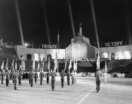 Large stadium celebration in support of the war effort. Troops carrying flags and facing a raised platform stage with an American eagle and a sign that reads "TRIBUTE TO VICTORY."