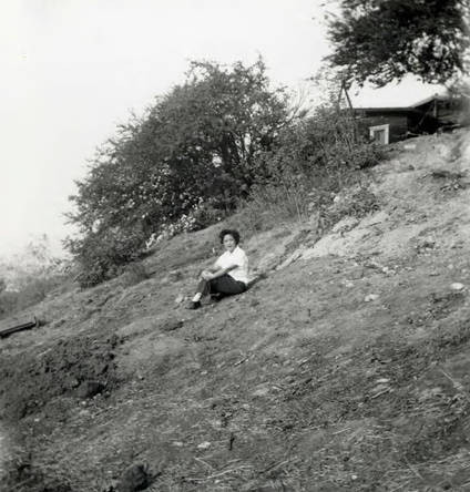 Woman seated on a hillside. Text on the back reads "Our lot the week before" (Spencer Chan Family)