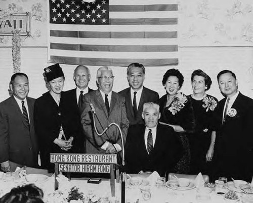Posed photo of a small group of people, including Gee Kee Ward, Senator Hiram Fong, a Caucasian couple, and Wilbert Woo, Mrs. Hong, Y.C. Hong, Mrs. Fong and David Chow before an American flag; taken at the Hong Kong Restaurant in Los Angeles