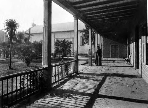 Exterior shot of the northerly wall of the Plaza Church and rectory. Palm treet and cross in yard in front of rectory. One Priest is standing in front of the rectory under the overhand of the rectory