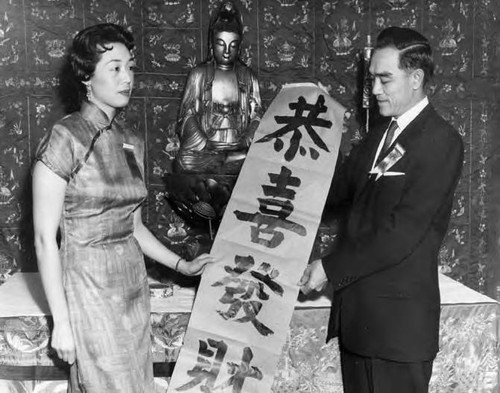 Man presents scroll featuring Chinese characters. In the top center is a seated Buddha figure