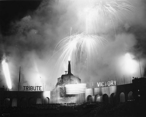 Large stadium celebration in support of the war effort. Large letters at the far top edge of the stadium read: "TRIBUTE TO VICTORY." Stadium is full of supporters and a firework display is directly above