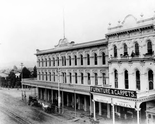 Pico House with balcony and stage in front and Merced Theater that says "Barker & Allen, Funiture & Carpets." Looking northeast with the Plaza in the background and trolley tracks on Main street