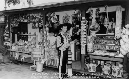 Print of a postcard showing Mario Valadez in Mexican dress standing in front of his Olvera Street curio shop. Shop was originally one of the 1932 Olympic cottages