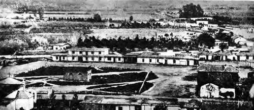 Photograph of brick waterworks in the center of the Plaza, the two story Lugo adobe and a corner of the Plaza Church