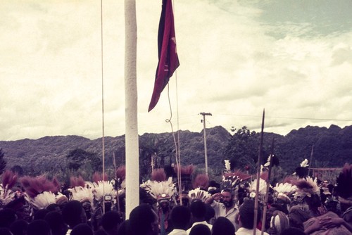 Newly adopted Papua New Guinea flag raised at Erave patrol post, watched by crowd including decorated men and saluting patrol officer