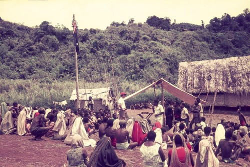 Erave patrol officer (in red cap), on patrol, addresses villagers, Australian flag hangs from pole (left)