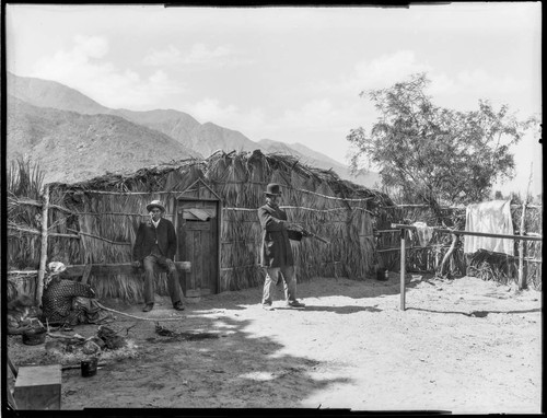 Woman, two men, one with rifle, outside wickiup