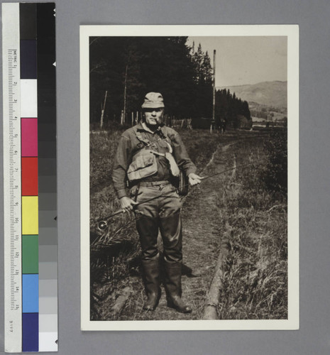 Edwin Powell Hubble, dressed in wading boots and fishing gear, holding a fishing rod, standing outdoors on a path in Rio Blanco Ranch, Colorado