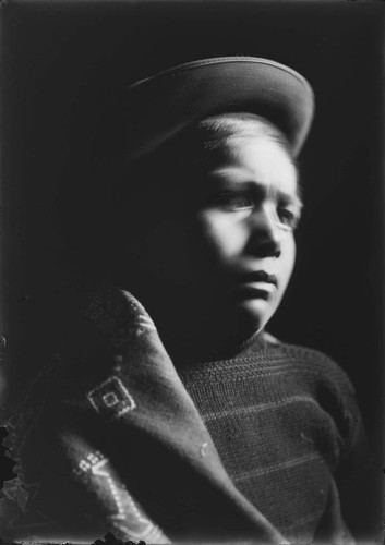 Portrait of Mountain Crow boy wearing felt hat, sweater, and blanket around shoulder