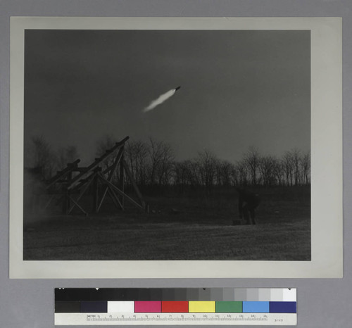 A rocket launched from wooden scaffolding, near a tree-lined field