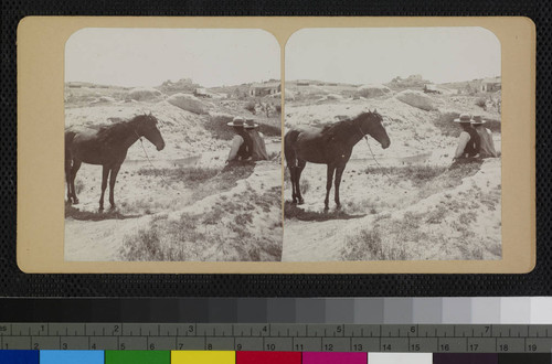 Two men and a horse next to a stream, with a covered wagon and house in the background