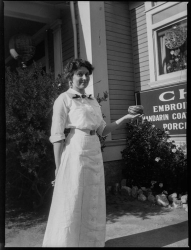 Grace Nicholson standing in front of her store and home at 46 N. Los Robles, Pasadena, holding a basket