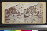 A Pueblo Indian stands near stairs leading to an unidentified pueblo on top of a mesa