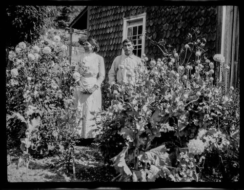 An unidentified Native American man and woman standing in front of a wood-shingled house, with garden