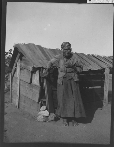 An unidentified Native American woman standing in front of wooden plank house