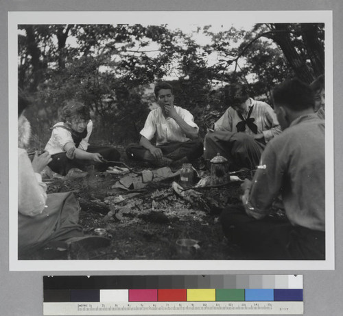 Edwin Powell Hubble with five unidentified companions, seated outdoors around a campfire, toasting bread