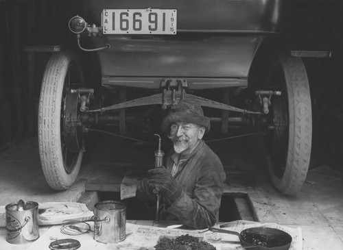 Ferdinand Ellerman standing in a pit below an automobile, with grease gun in hand