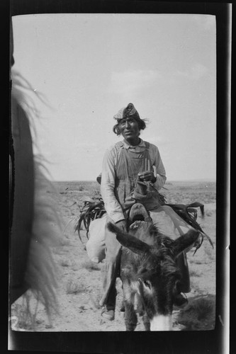 Hopi man riding a burro carrying a load - possibly corn