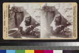 A Hopi woman is crouched near a pile of corn and a pottery bowl, in an unidentified village