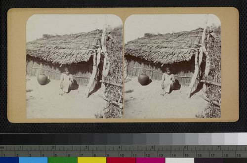 A Cahuilla young woman sitting outside a brush house, with a pottery jar next to her