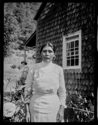 An unidentified Native American woman standing in front of a wood-shingled house, with Grace Nicholson standing in background