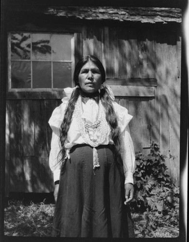 An unidentified Native American woman wearing necklace of dentalia, standing in front of house