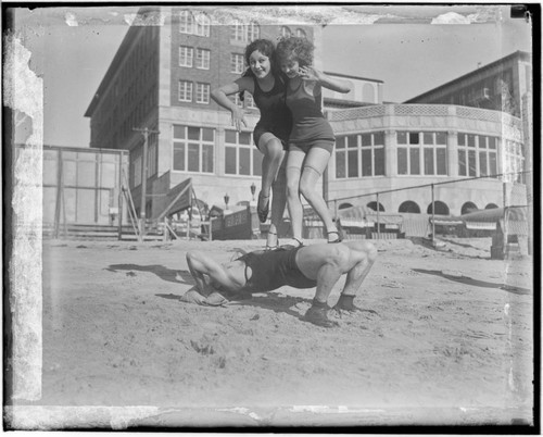 Two women standing on the torso of Henry Steinborn in front of Club Casa del Mar