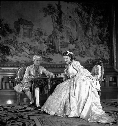 Couple in period costume seated at a small table in the library of the Huntington residence with a Beauvais tapestry in the background