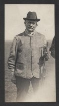 Man in hat and vest, standing in a field with a rifle