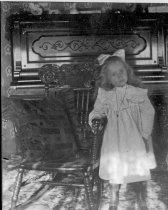 Young girl in ruffled dress next to rocking chair and piano, c. 1912