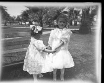 Two children in dresses, holding hands in park in front of palm trees