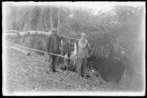 Two men in woods, holding poles with caught fish