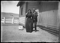 Couple posing arm in arm in front of farm buildings, c. 1906