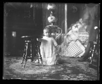 Infant in dress, standing next to stool