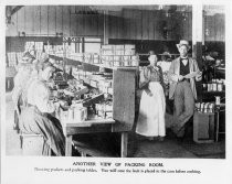 "Another view of packing room. Showing packers and packing tables. You will note the fruit is placed in the cans before cooking."