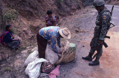 A soldier inspecting a Mayan man's basket in search of subversive materials, Chichicastenango, 1982