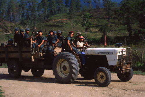 Group of armed Contra soldiers sit on a wagon hauled by a white tractor, Nicaragua, 1983